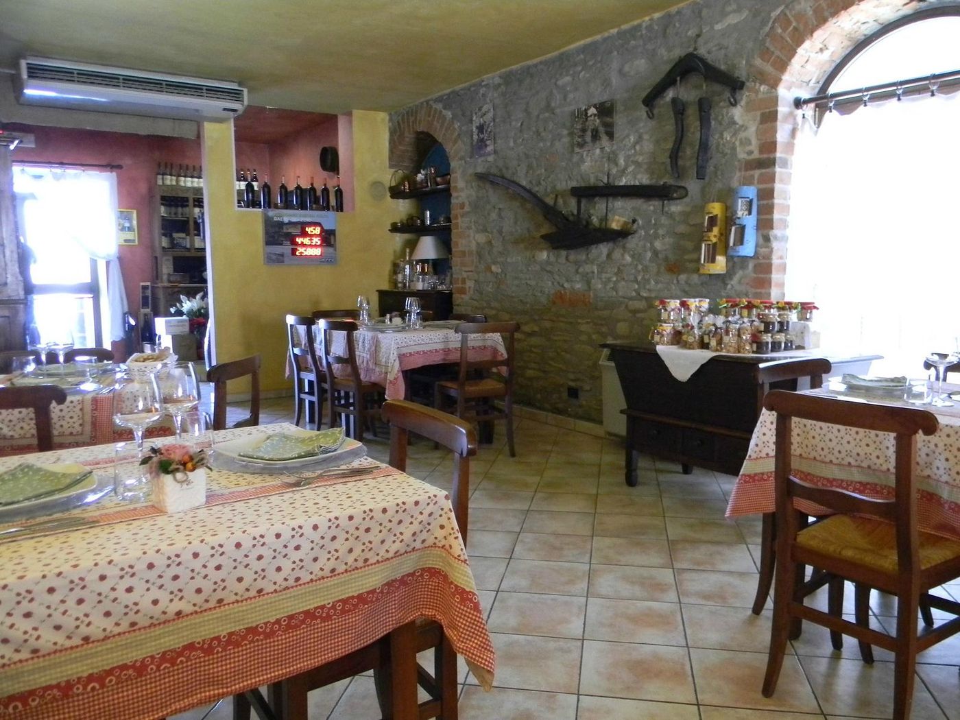 La sala dell'osteria: muro in pietra, attrezzi agricoli appesi, tovaglie a quadri